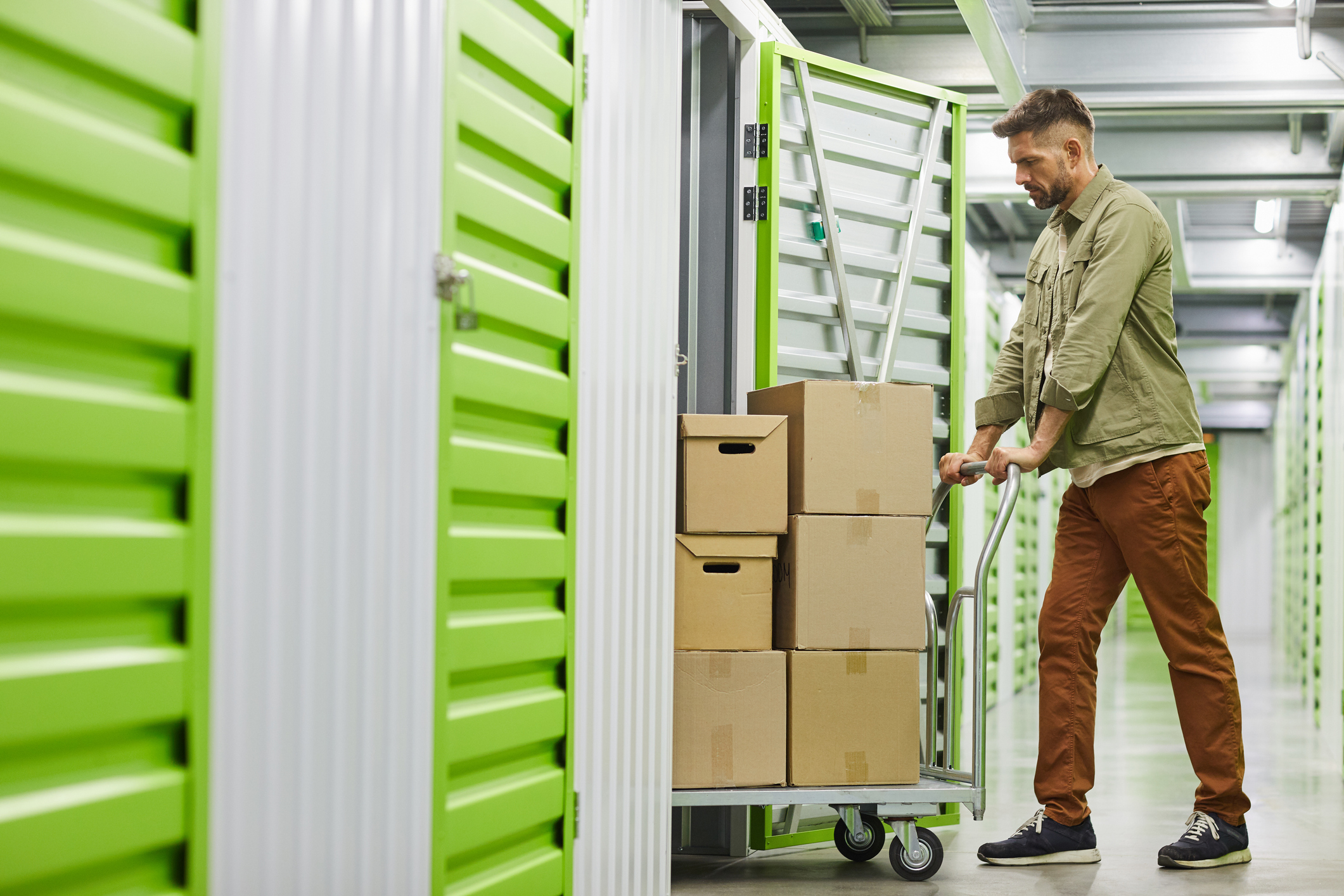 Full length side view at handsome bearded man loading cart with cardboard boxes into self storage unit, copy space